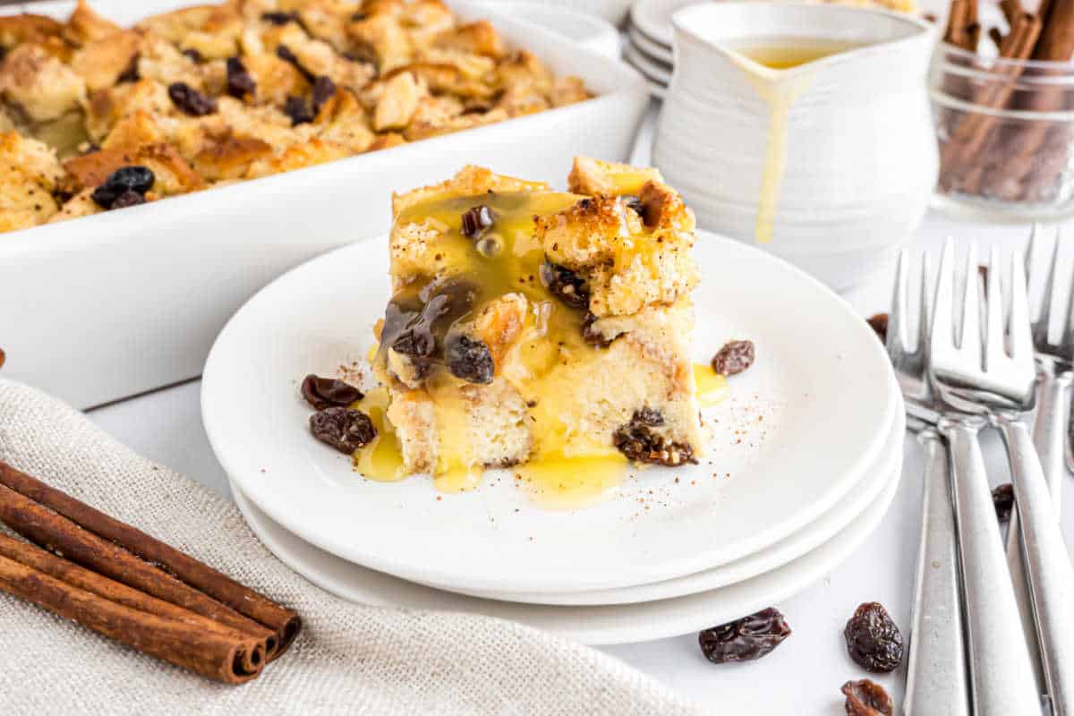 Two slices of bread pudding made from a classic bread pudding recipe with raisins, topped with sauce, are served on a plate with forks and cinnamon sticks nearby.