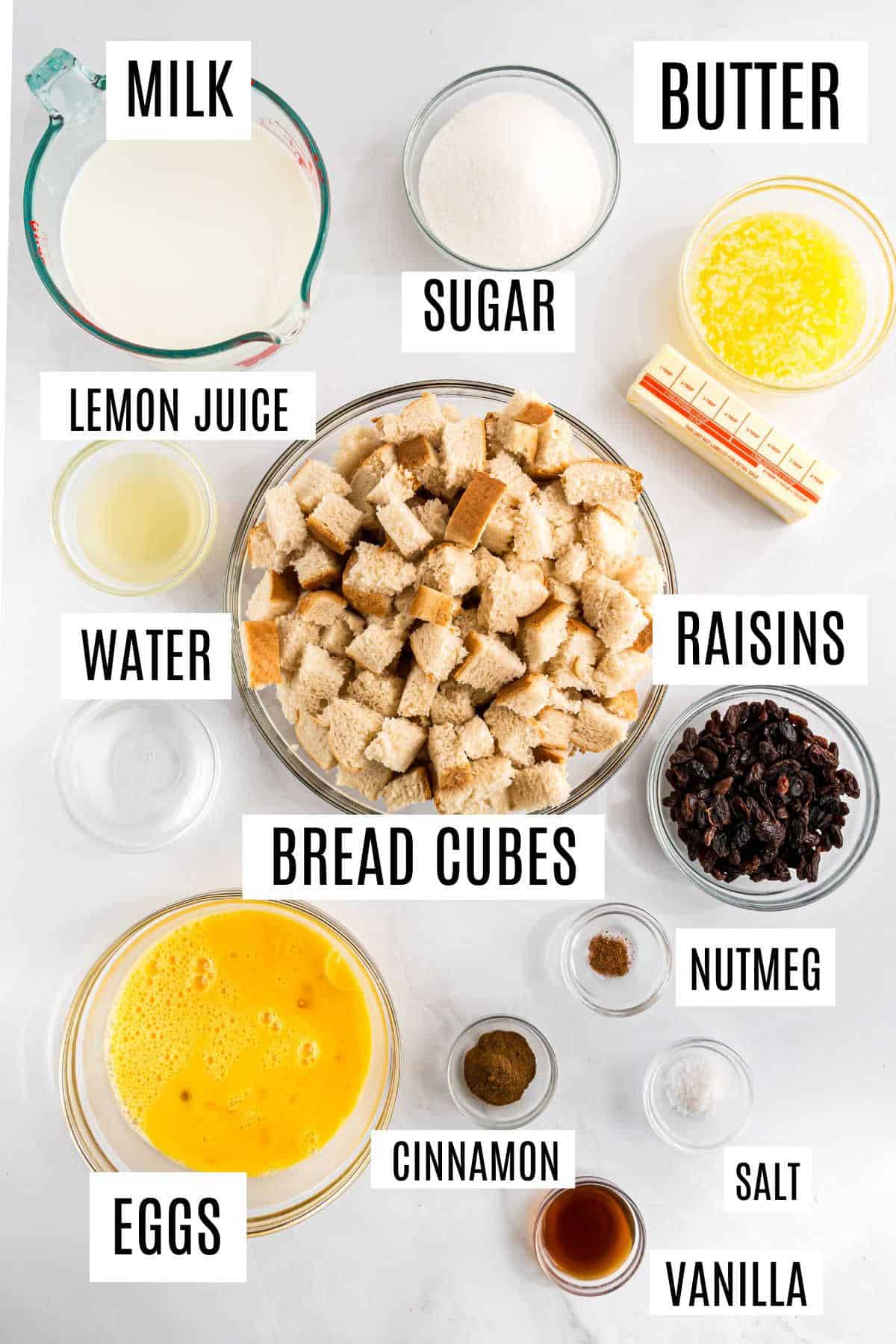 Overhead view of measured bread pudding recipe ingredients labeled: milk, eggs, bread cubes, raisins, sugar, butter, spices.