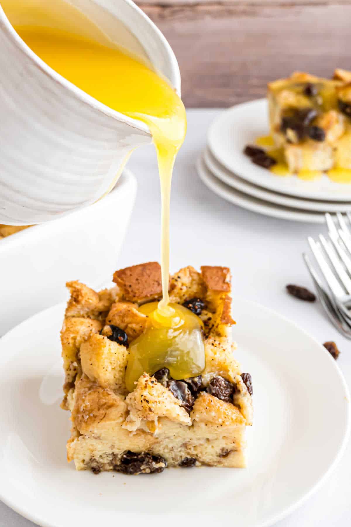A slice of bread pudding from our delicious bread pudding recipe sits on a plate, rich sauce being poured over it, with more slices tempting in the background.