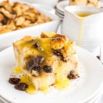A slice of bread pudding made from a classic bread pudding recipe with raisins and vanilla sauce on a white plate, with sauce pouring in the background.
