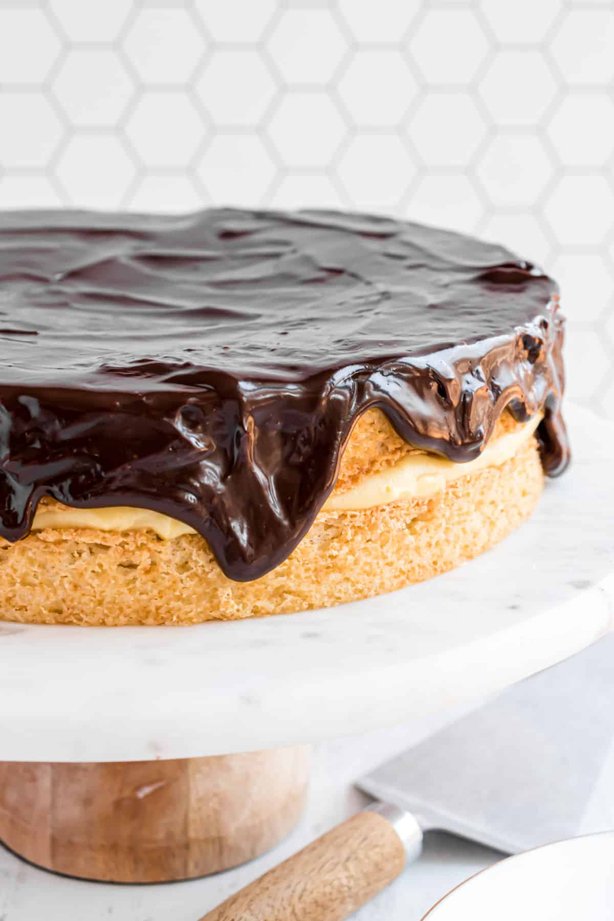 Boston cream pie on white cake plate with ganache dripping down sides.