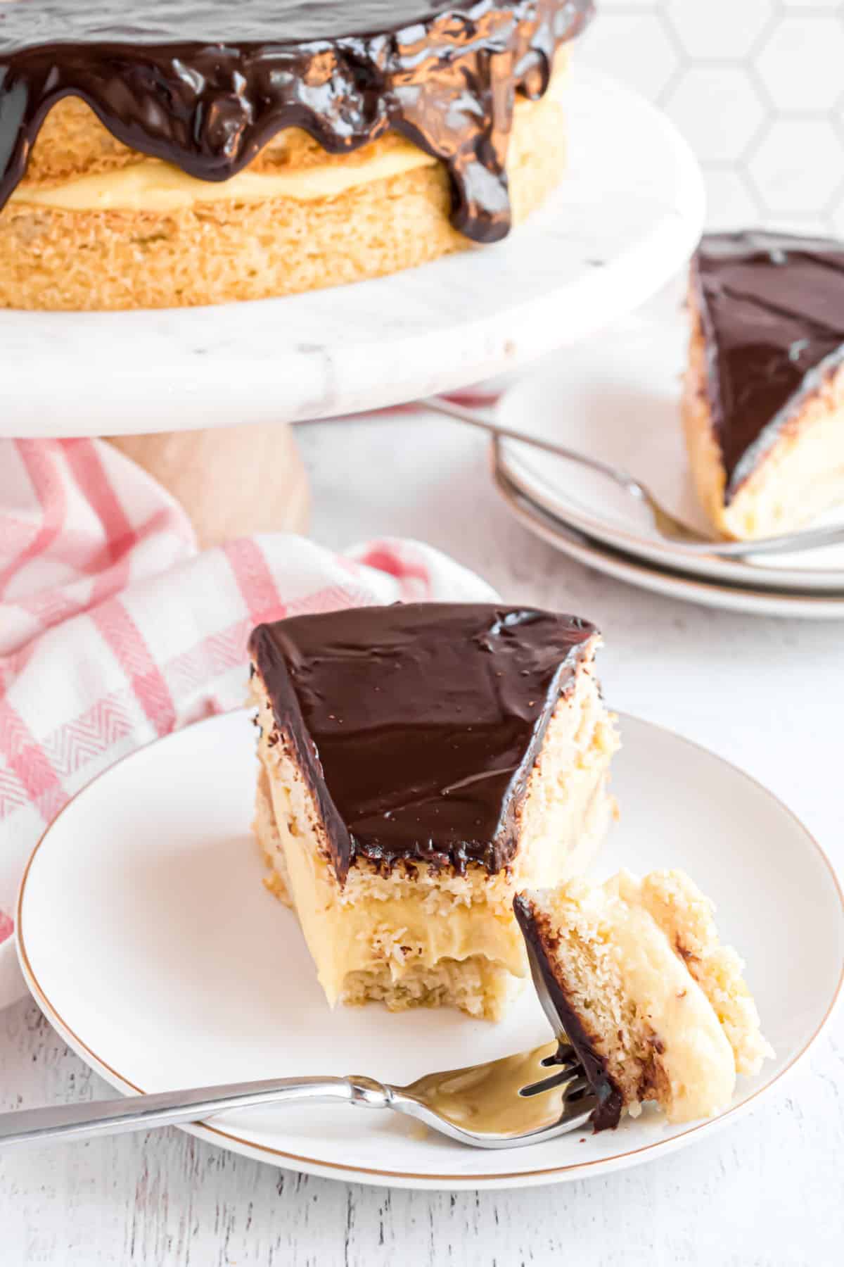 Slice of boston cream pie on a white plate with a fork and a bite taken.