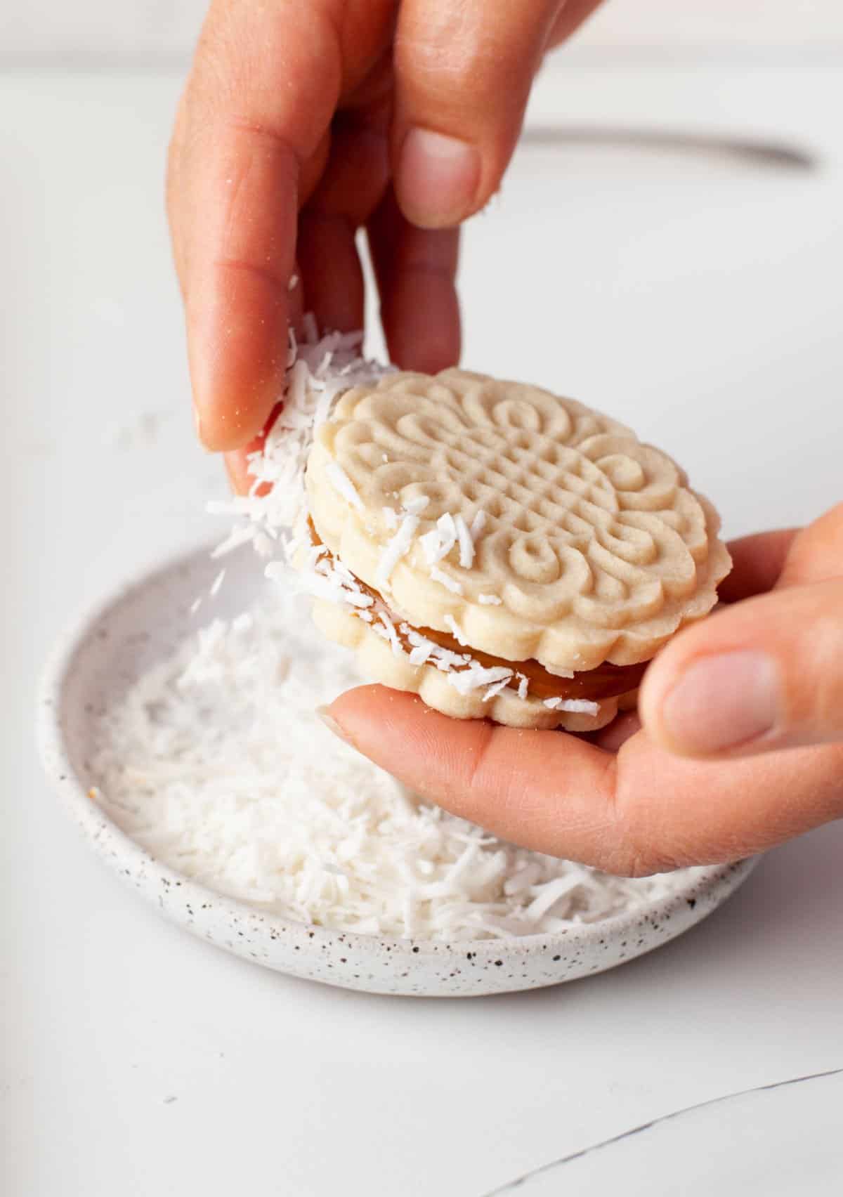 Alfajores cookie assembled with coconut being pressed to sides of cookie.