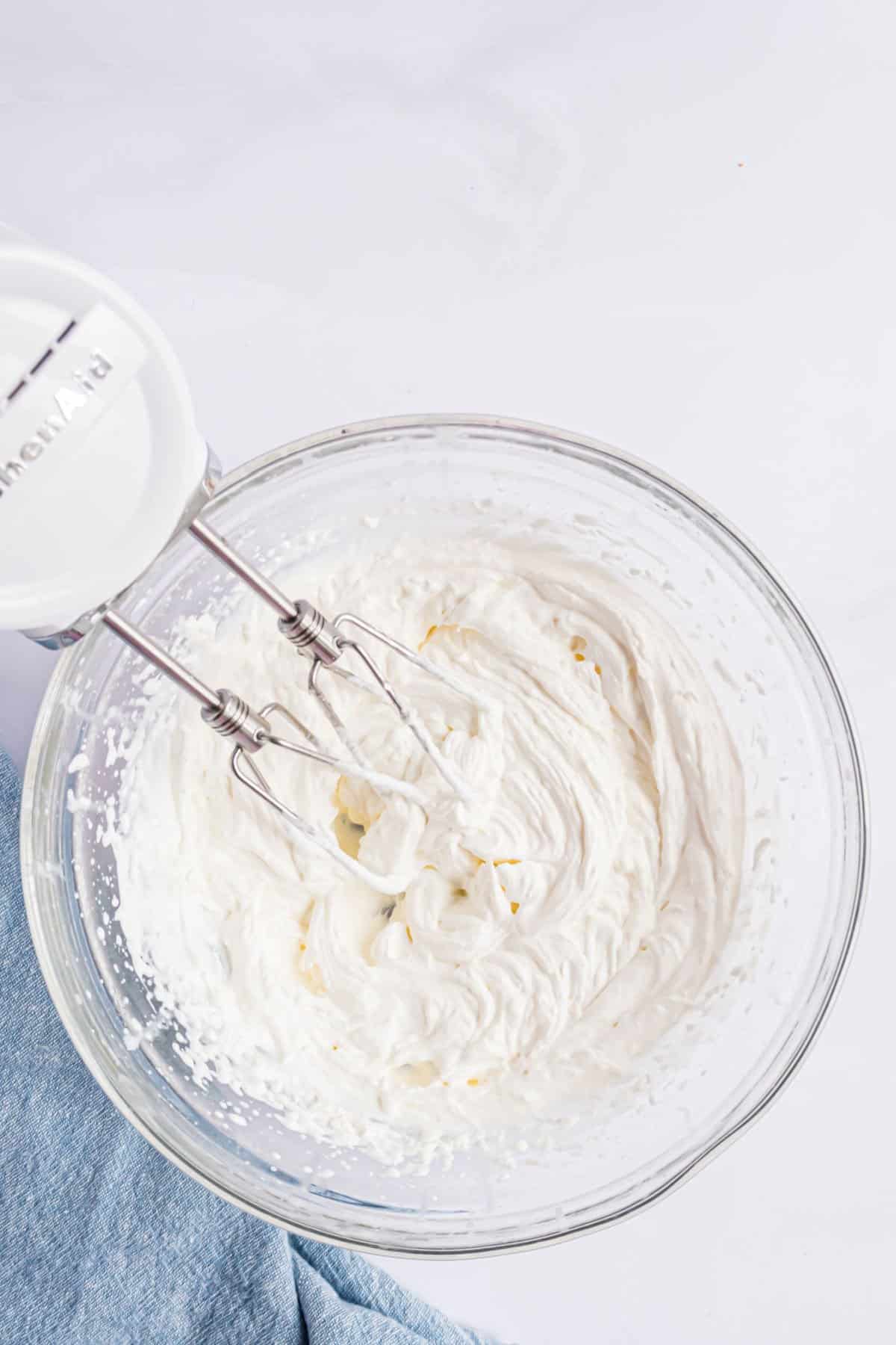 Stabilized whipped cream in clear glass mixing bowl with hand mixer.
