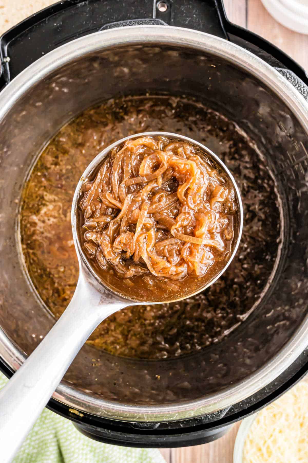 A ladle holding caramelized onions hovers over a pot filled with rich, brown French onion soup.