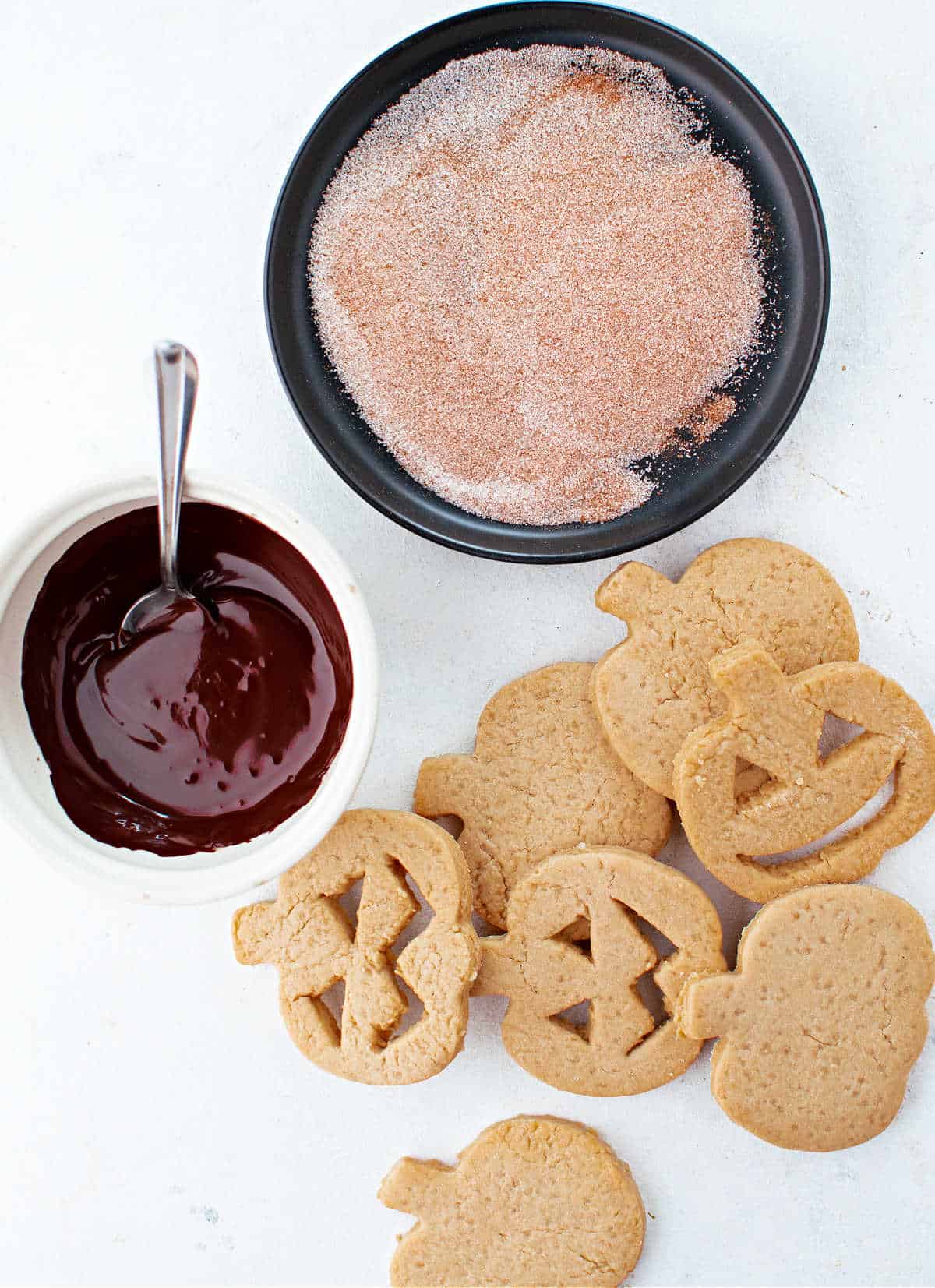 Melted chocolate and cinnamon sugar mix in bowls with pumpkin cut out cookies before assembly.