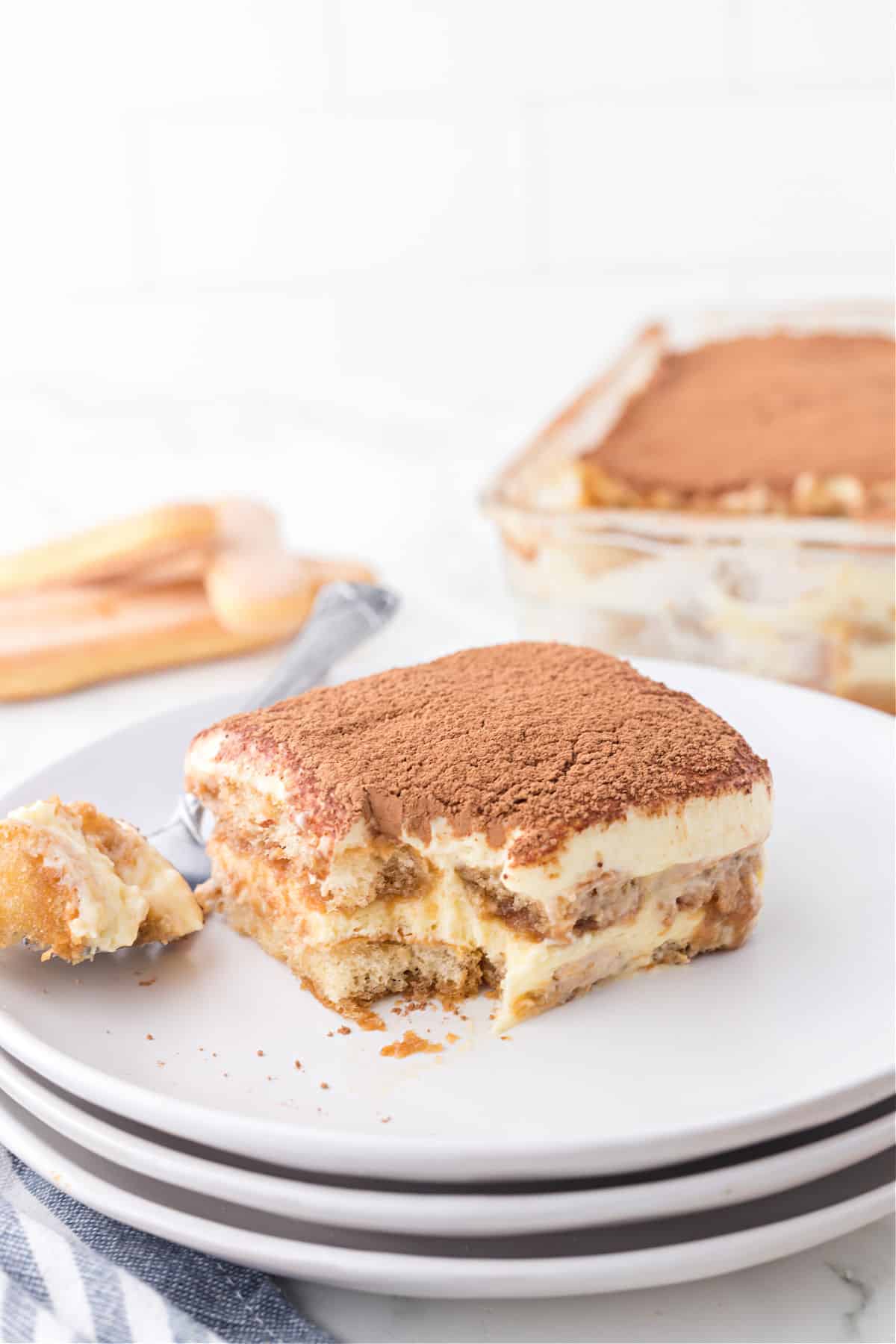 Stack of white plates with a slice of tiramisu and a bite taken out of it.