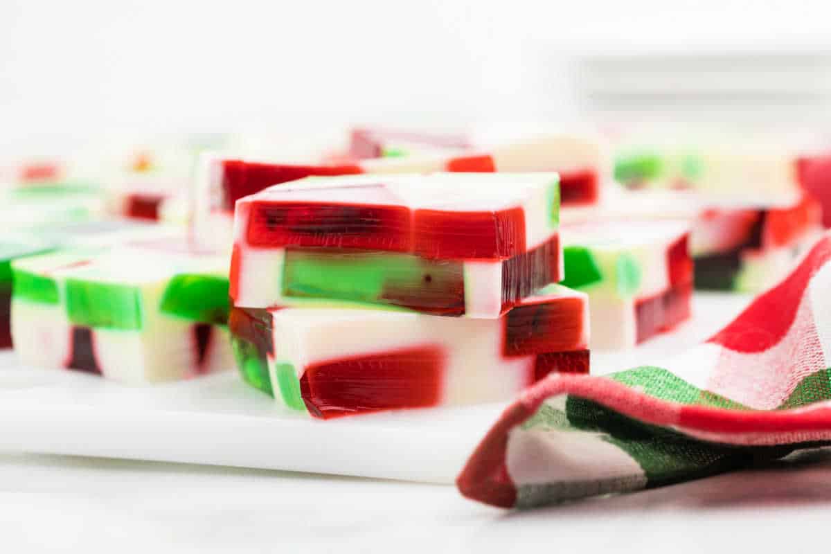 Red, green, and white layered holiday Jello squares, inspired by broken glass Jello, are stacked on a white surface with a checkered cloth nearby.