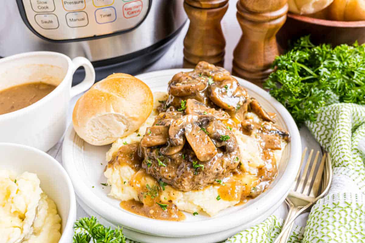 Instant Pot Salisbury steak with mushroom gravy over mashed potatoes, served with a roll, is presented in front of an Instant Pot for a hearty, homestyle meal.