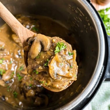 A wooden spoon lifts creamy mushroom gravy from a pot of instant pot salisbury steak, garnished with fresh parsley.