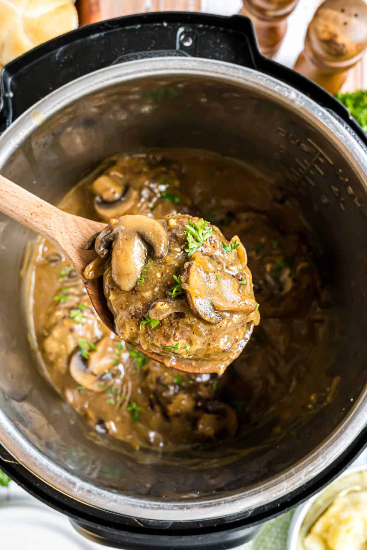A wooden spoon lifts tender Instant Pot Salisbury steak with mushrooms and rich gravy from an Instant Pot.