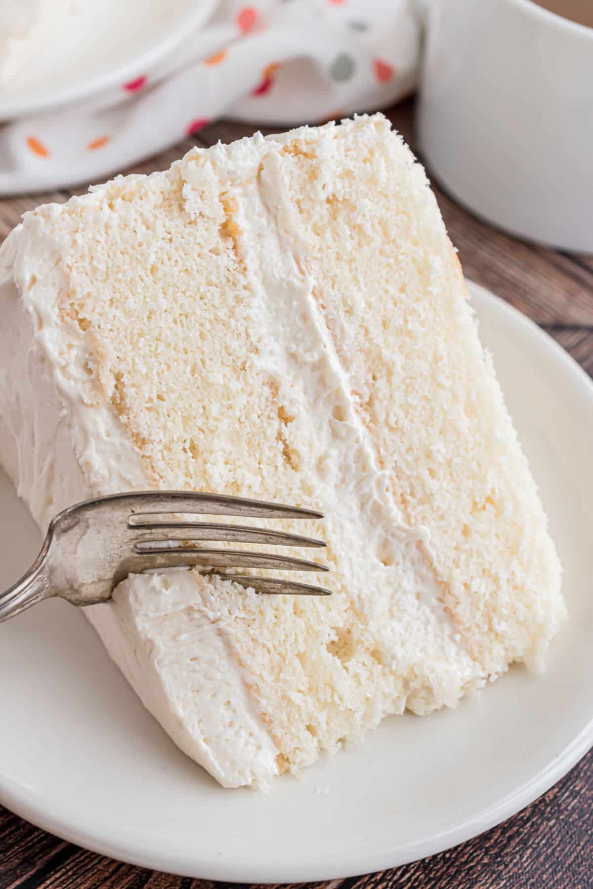 Slice of white layer cake with vanilla frosting on a plate with a fork.