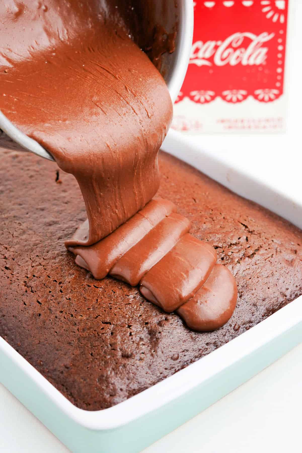 Chocolate frosting being poured over a chocolate cake in a 13x9 pan.