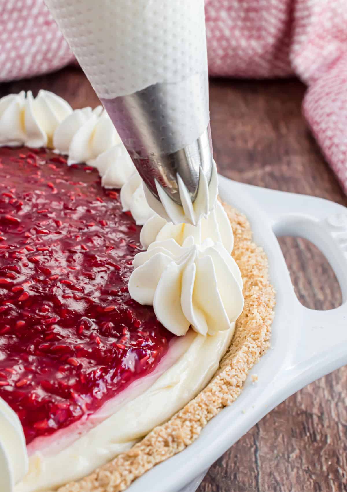 Whipped cream being piped onto a raspberry cheesecake.