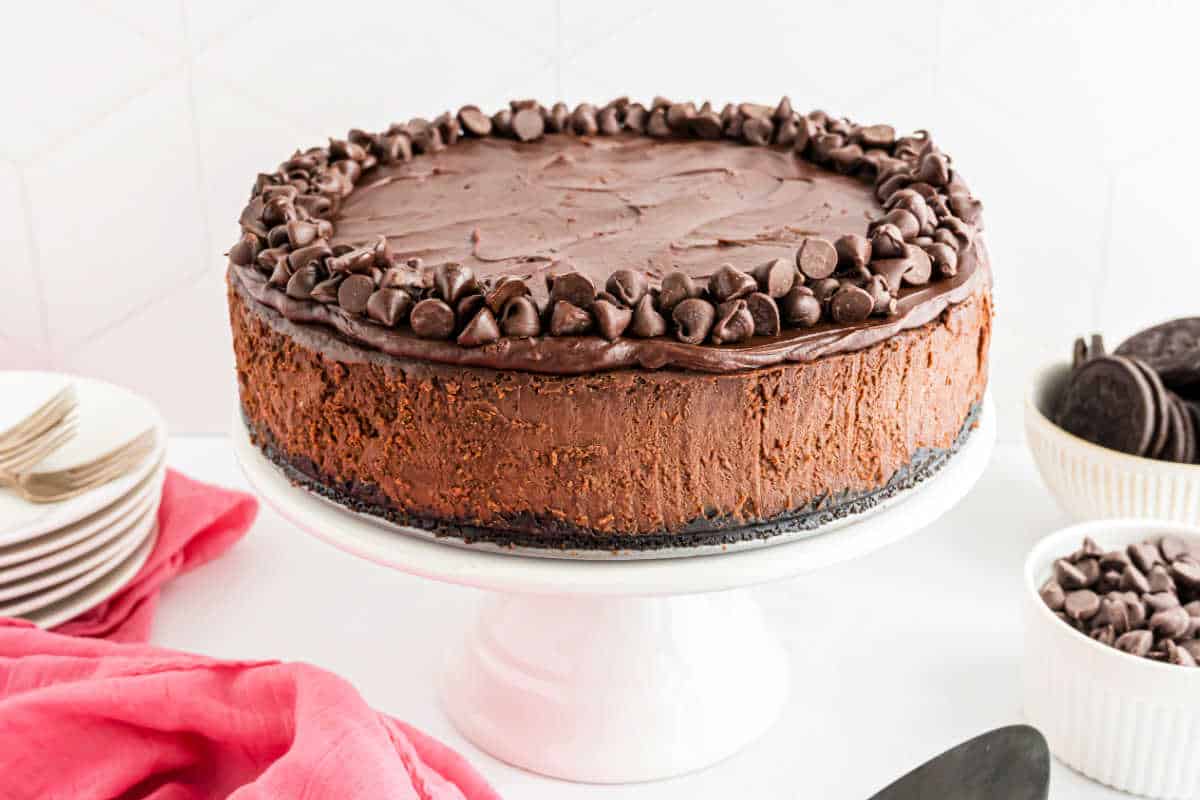 Chocolate cheesecake topped with chocolate chips sits elegantly on a white cake stand, surrounded by plates and cookies nearby.