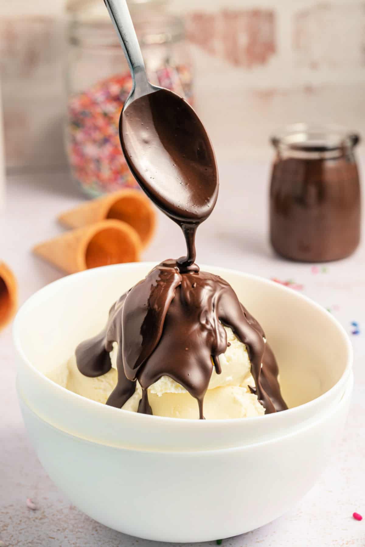 Chocolate sauce being drizzled over a bowl of vanilla ice cream with a spoon.