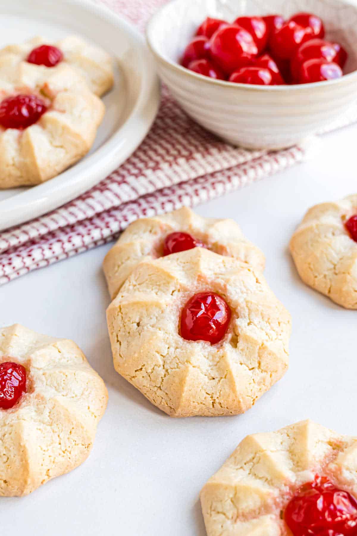 Amaretti cookies topped with red cherries are arranged on a white surface, with a bowl of cherries nearby.