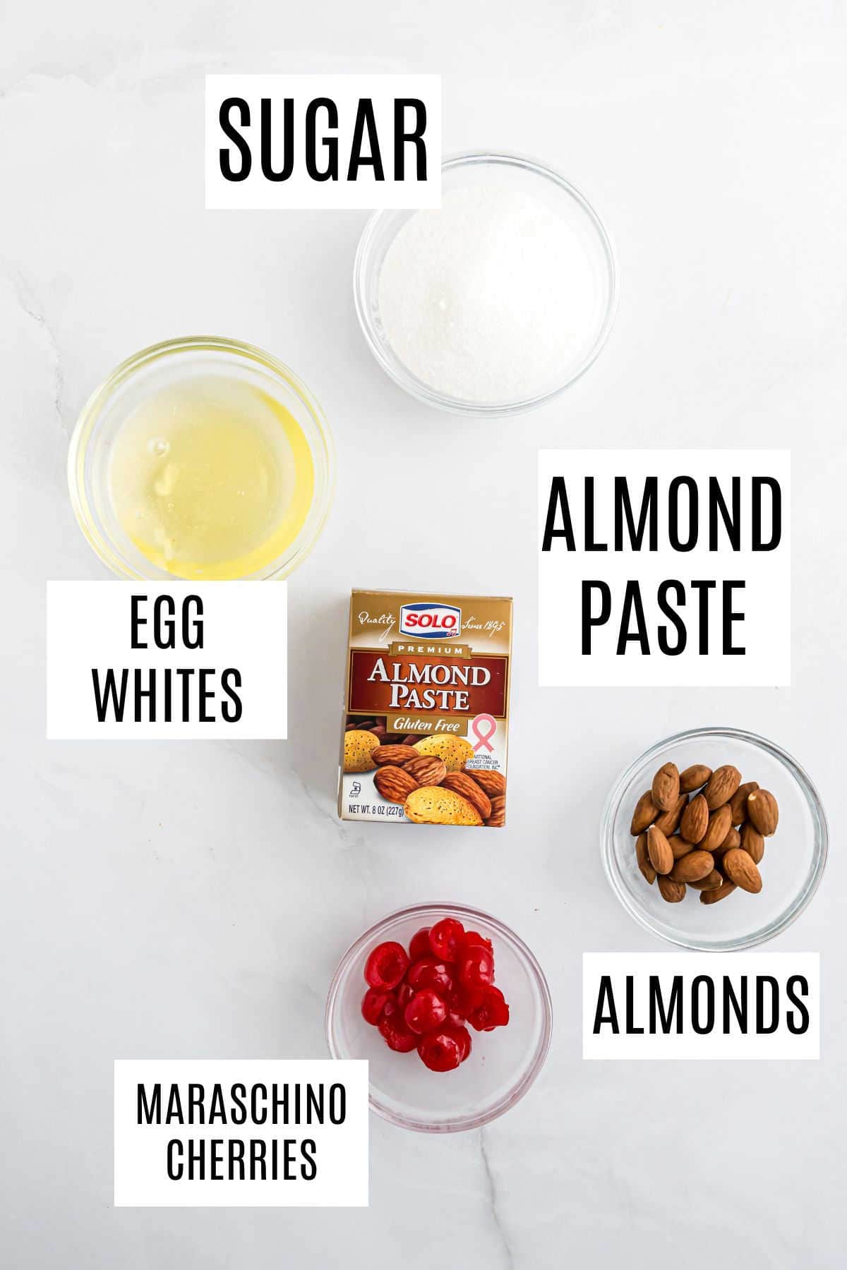Six ingredients for baking amaretti cookies: sugar, egg whites, almond paste, almonds, and maraschino cherries arranged on a white surface.