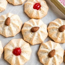 Round amaretti cookies topped with whole almonds or red cherries rest on parchment paper.