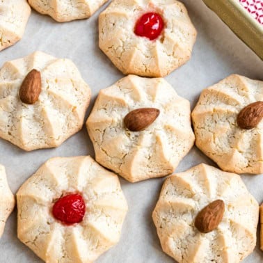 Round amaretti cookies topped with whole almonds or red cherries rest on parchment paper.
