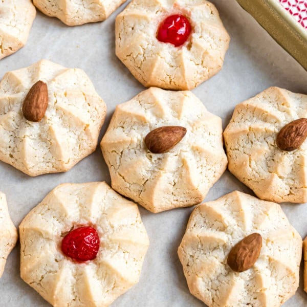 Round amaretti cookies topped with whole almonds or red cherries rest on parchment paper.