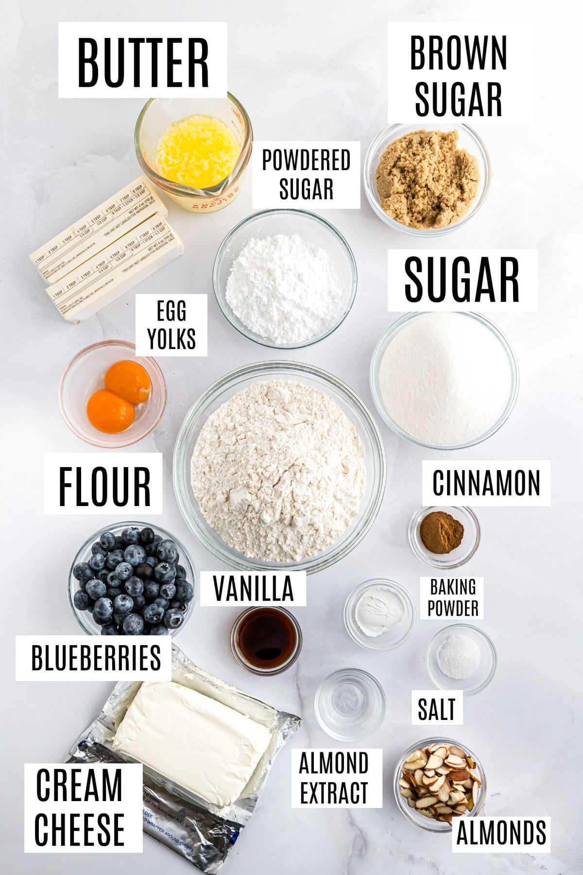Top-down view of ingredients for blueberry coffee cake cookies in bowls, labeled: butter, sugars, flour, egg yolks, cinnamon, blueberries, and more.