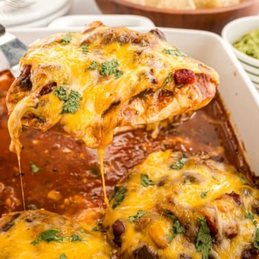 A cheesy baked salsa chicken breast with sauce and cilantro is being lifted from a casserole dish.