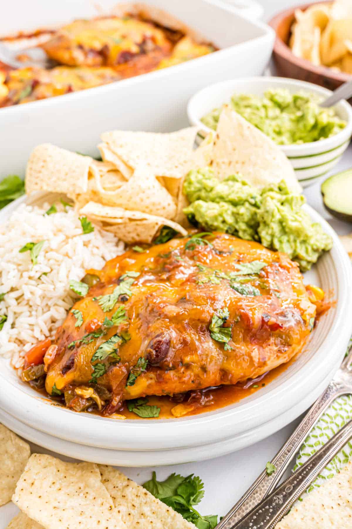 A plate with cheesy enchiladas, baked salsa chicken, white rice, tortilla chips, and guacamole, all garnished with fresh cilantro.