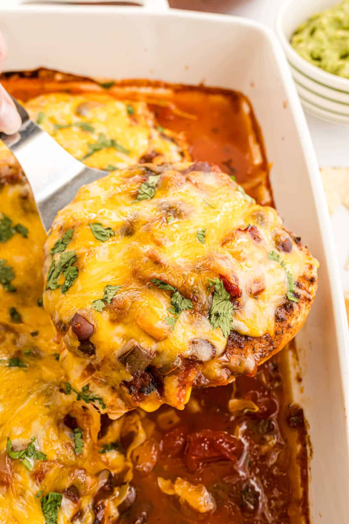 A spatula lifts a cheesy, baked salsa chicken enchilada from a casserole dish filled with sauce and melted cheese.