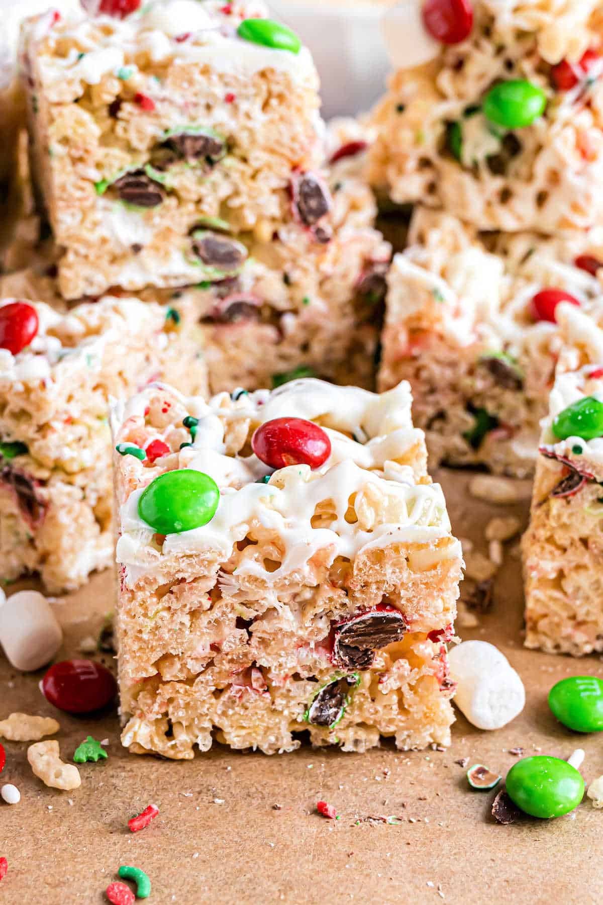 Festive holiday rice krispie treat squares, adorned with a white drizzle, are mixed with red and green candies and chocolate chips, all beautifully displayed on a rustic brown paper surface.