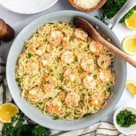 Large skillet of shrimp scampi pasta with lemon slices, parsley, and grated cheese, garnished with fresh herbs.