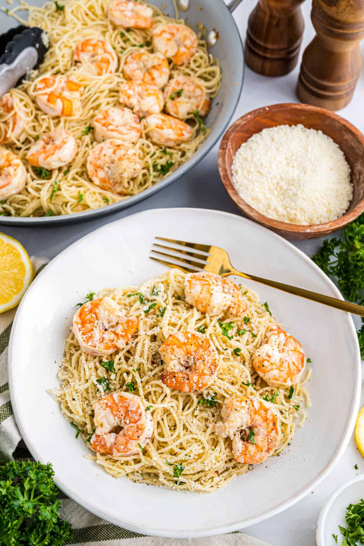 A plate of shrimp pasta, reminiscent of shrimp scampi, with grated cheese and parsley, sits in front of more pasta, cheese, and seasonings in the background.
