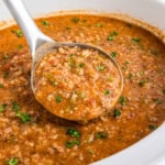A ladle serves a hearty stuffed pepper soup with beef and vegetables, topped with herbs in a white pot.