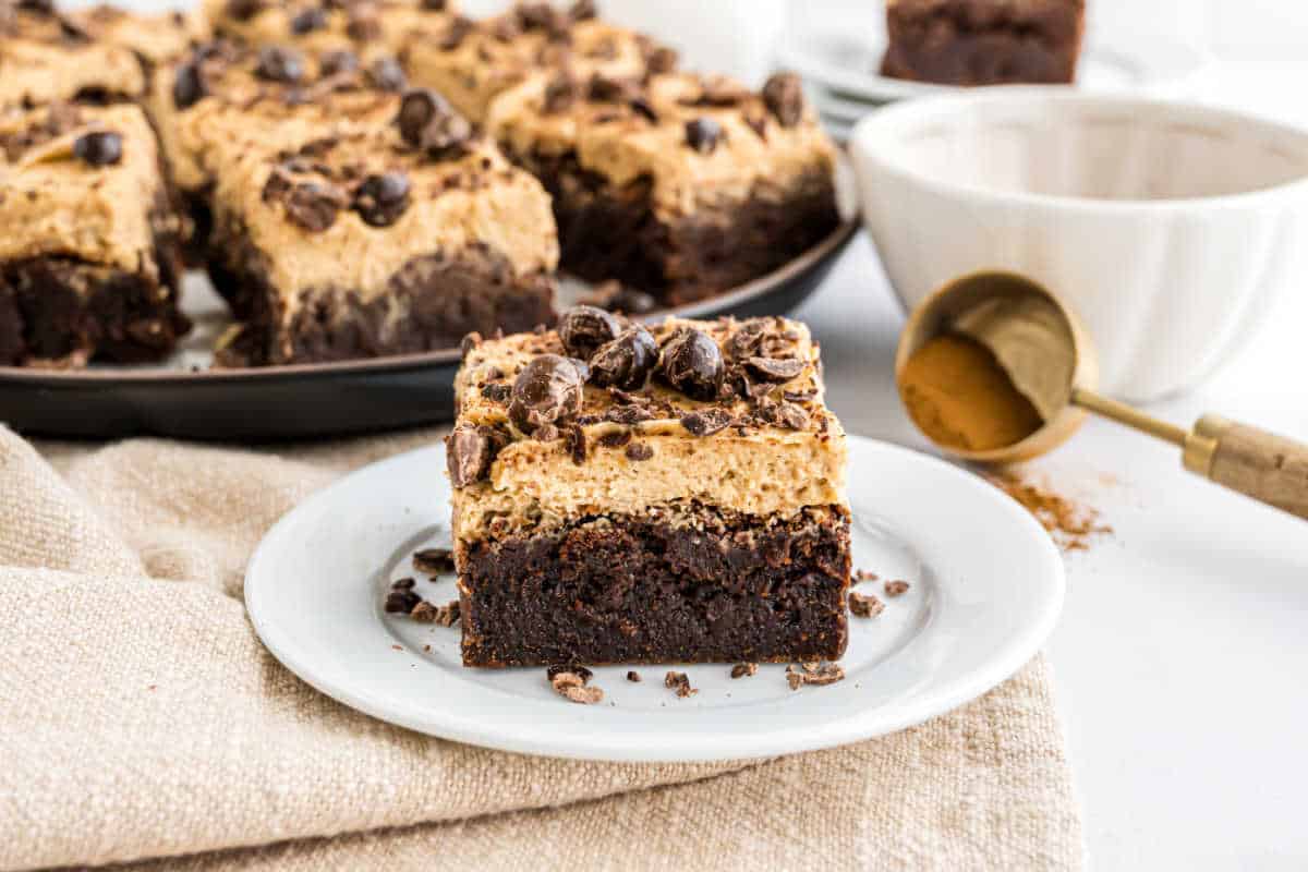 A square espresso brownie with creamy topping and chocolate bits on a white plate, with more brownies behind.