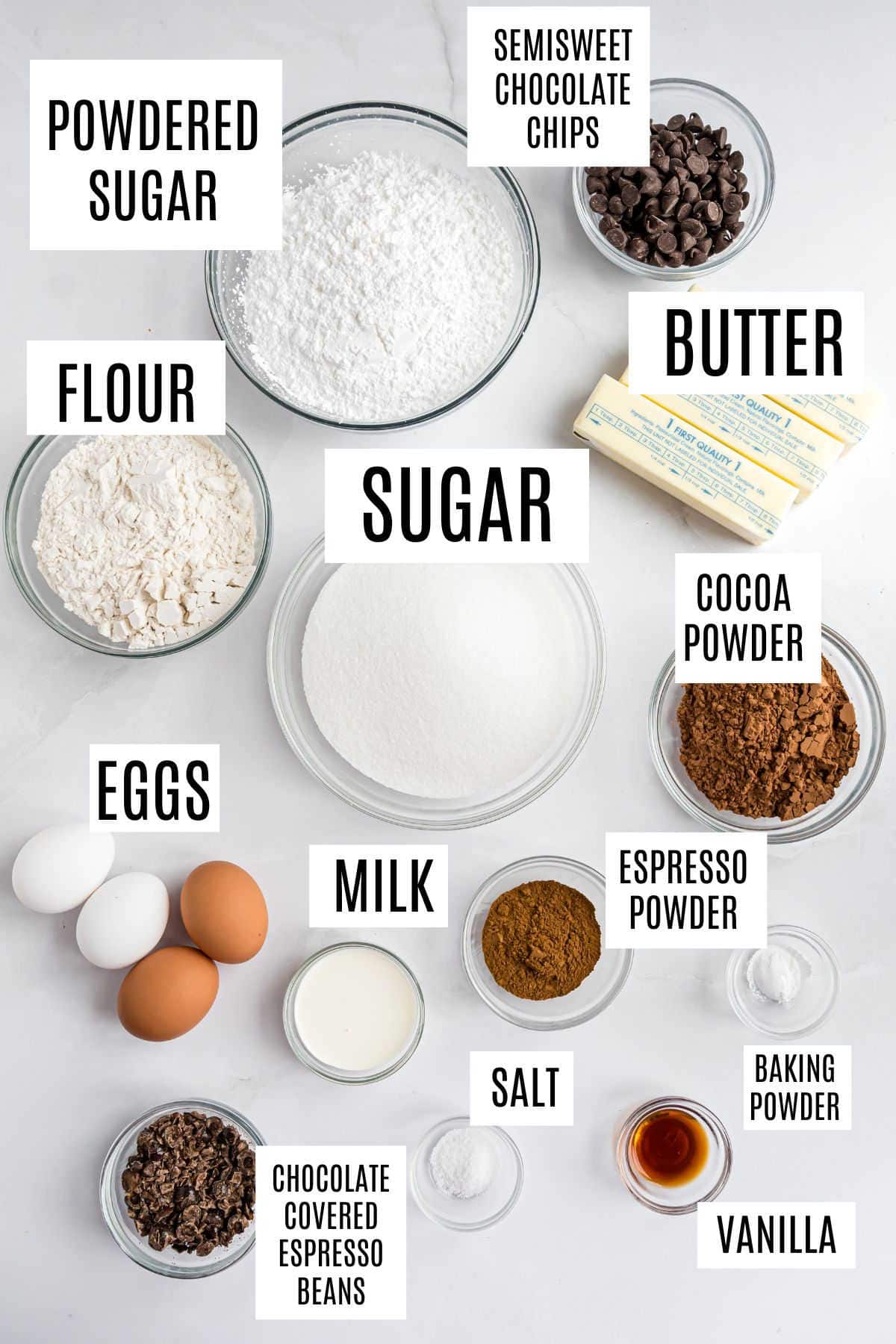 Baking ingredients labeled in bowls on a white surface, including sugar, flour, eggs, cocoa, and chocolate chips—perfect for making rich espresso brownies.