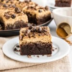 An espresso brownie topped with creamy frosting and chocolate pieces on a white plate, with more espresso brownies in the background.