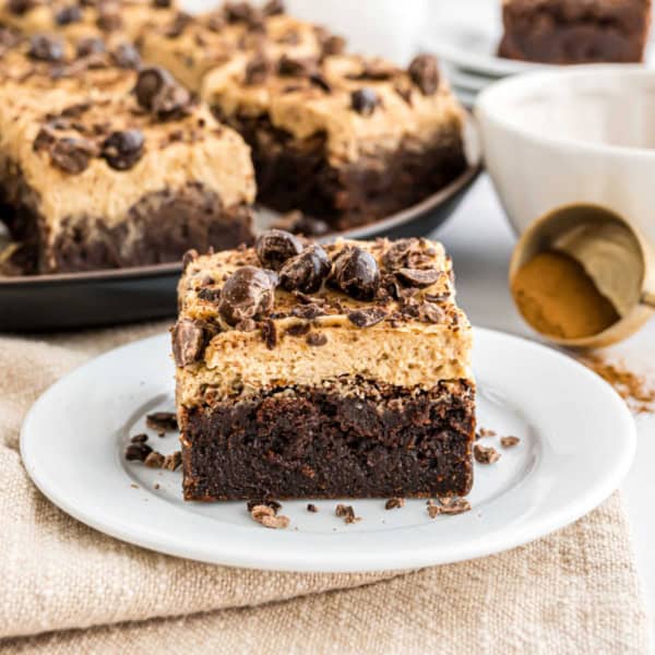 An espresso brownie topped with creamy frosting and chocolate pieces on a white plate, with more espresso brownies in the background.