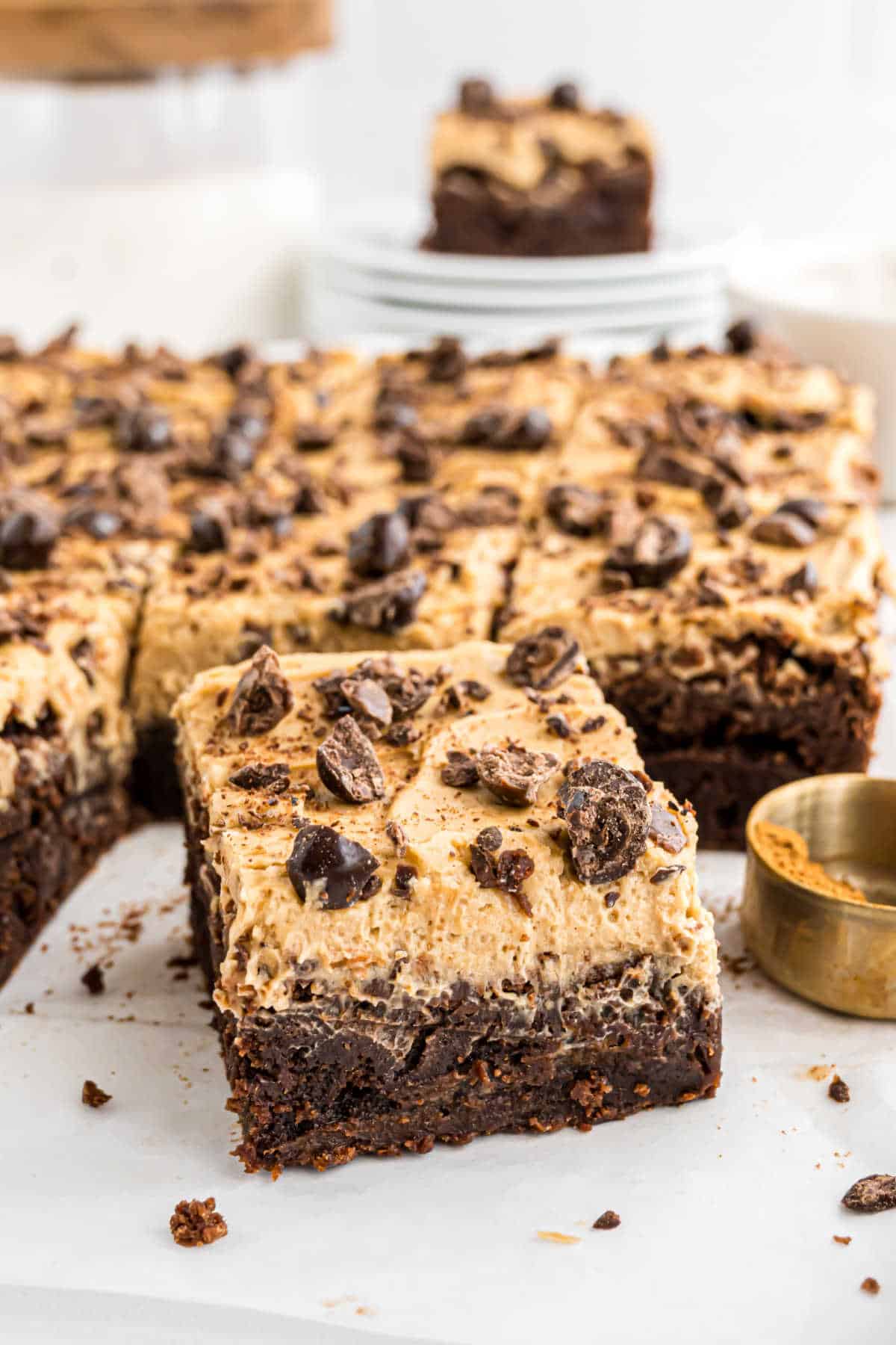A close-up of frosted espresso brownies topped with chocolate chunks, with one piece pulled forward on parchment paper.