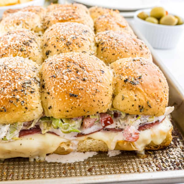 Savor the delightful Italian grinder sliders: baked sliders topped with herbs and cheese, filled generously with deli meats, lettuce, and perfectly melted cheese spilling from the sides.