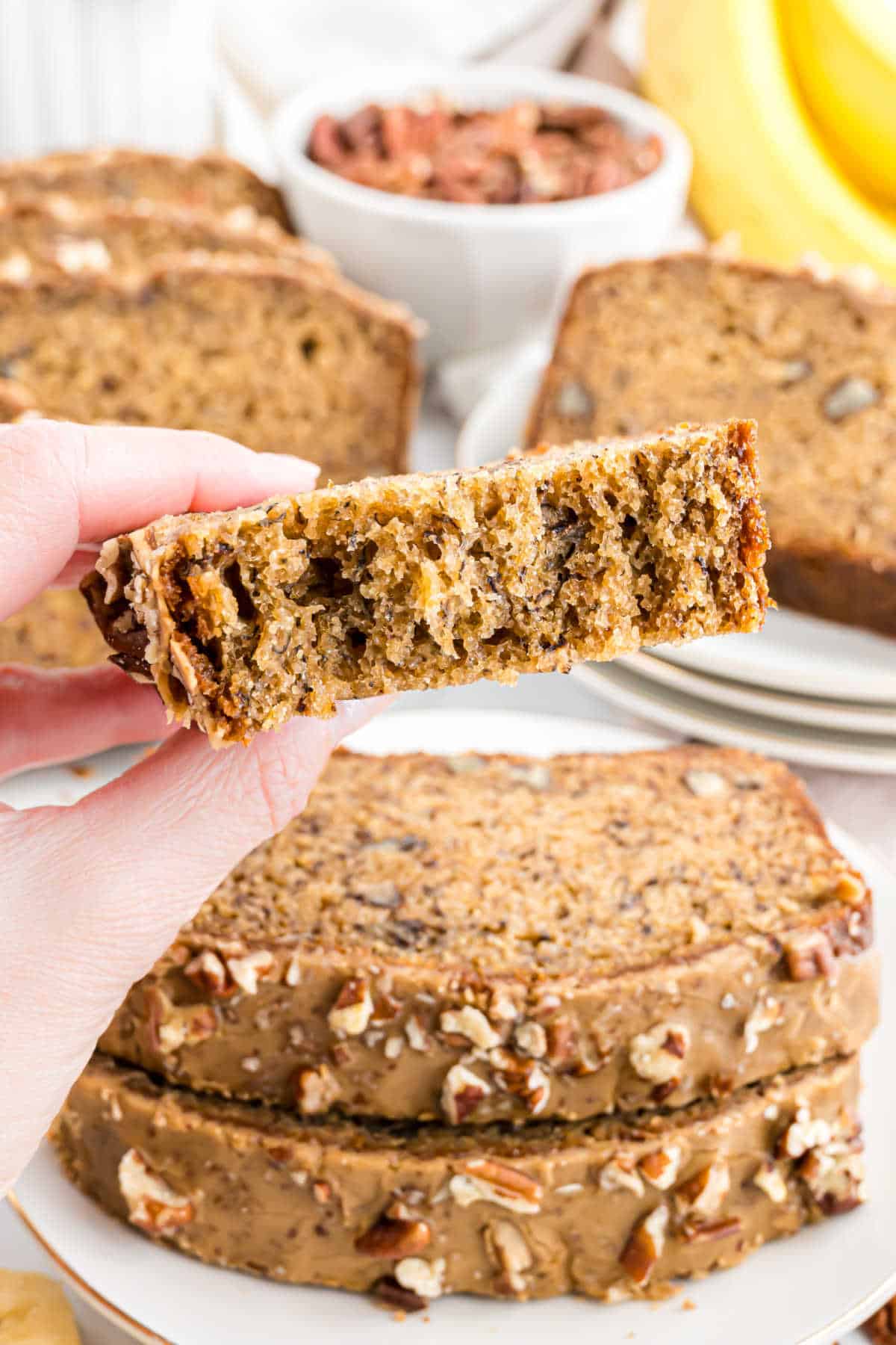 A hand holds a slice of maple pecan banana bread with nuts, with more slices and bananas in the background.