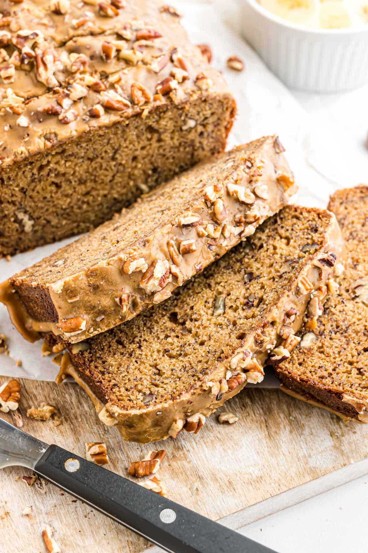 Sliced loaf of maple pecan banana bread with icing and chopped pecans on a wooden cutting board.
