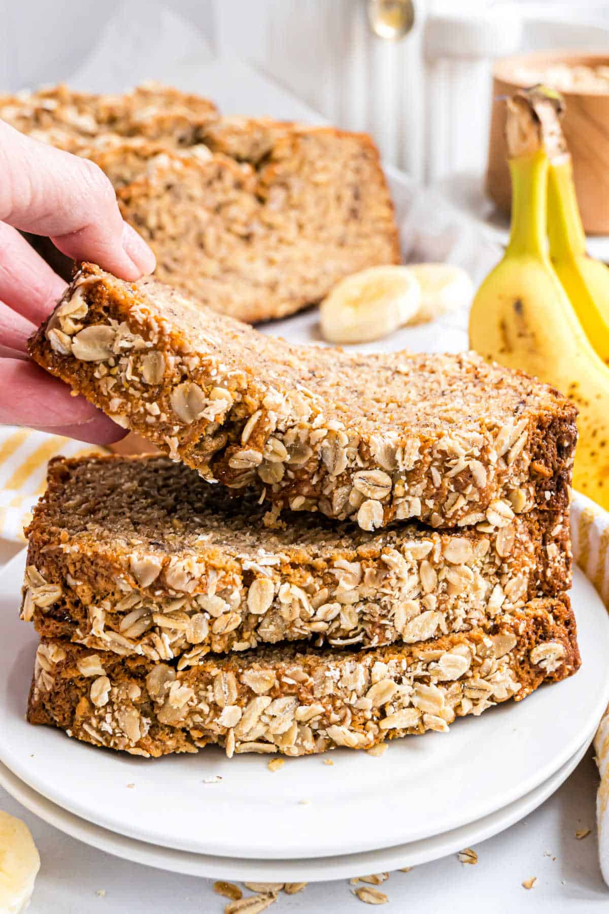 A hand lifts a slice of oatmeal banana bread from a stack on a white plate, with bananas in the background.