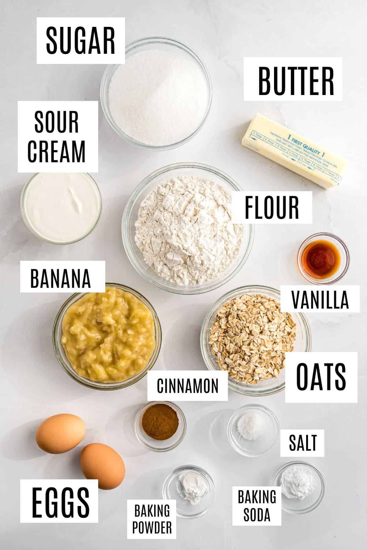 Overhead view of labeled ingredients in bowls for oatmeal banana bread: sugar, butter, flour, banana, sour cream, eggs, oats, cinnamon, and more.