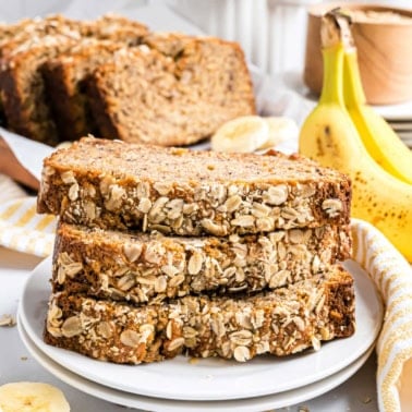 Three slices of oatmeal banana bread, oat-crusted and golden, are stacked on a plate, with fresh bananas and more bread in the background.