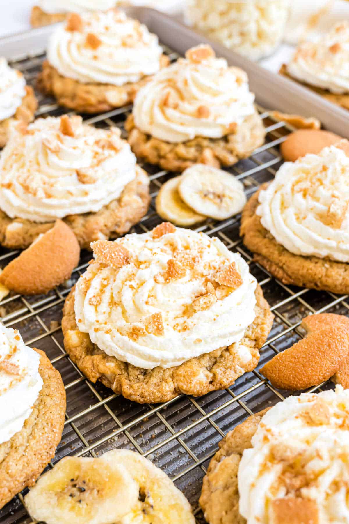 Cookies topped with swirled cream frosting and crumbled toppings, surrounded by banana chips and wafers on a cooling rack.