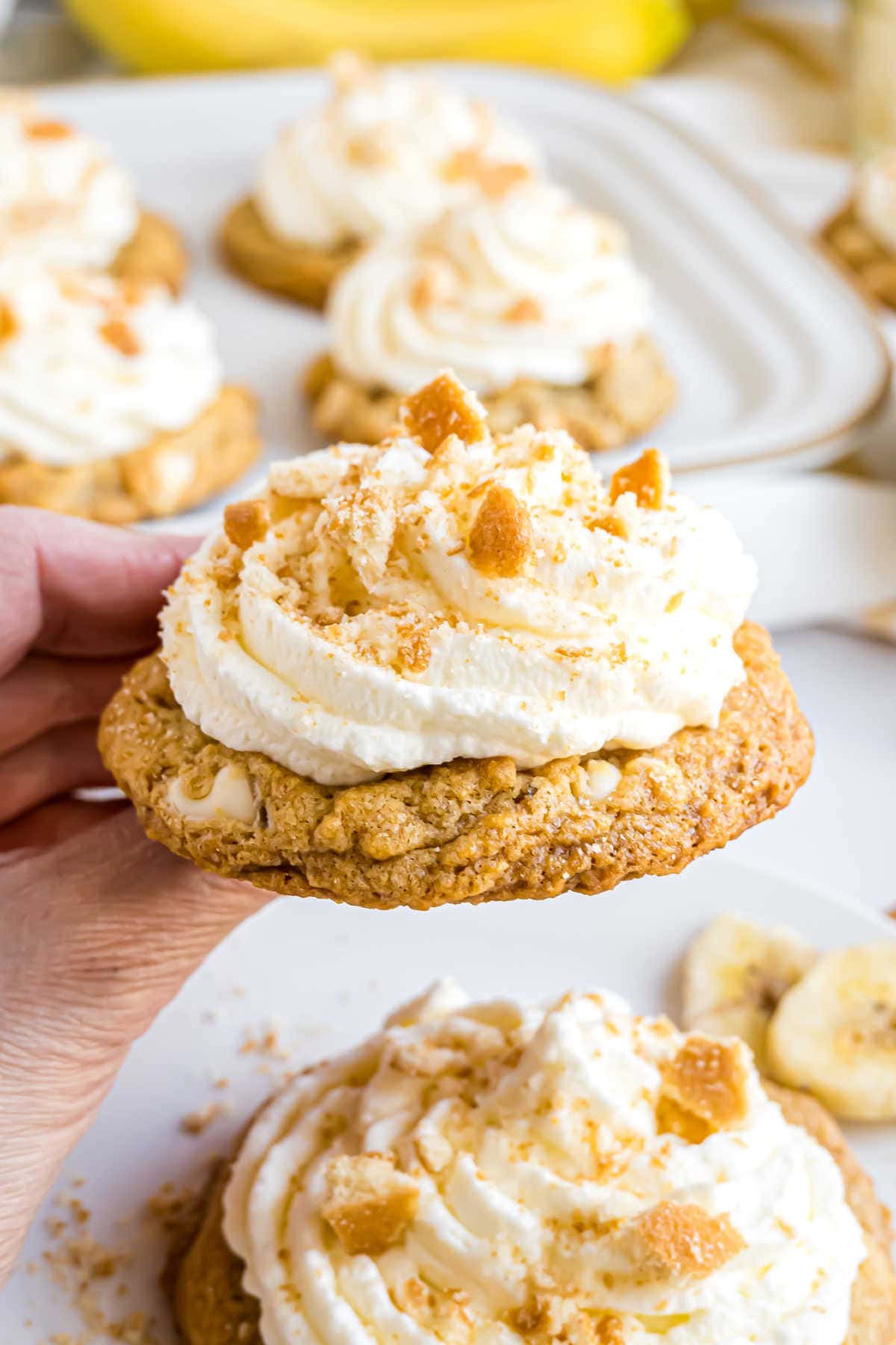 A hand holding a cookie topped with whipped cream and crumbled pieces, with more cookies on a tray in the background.