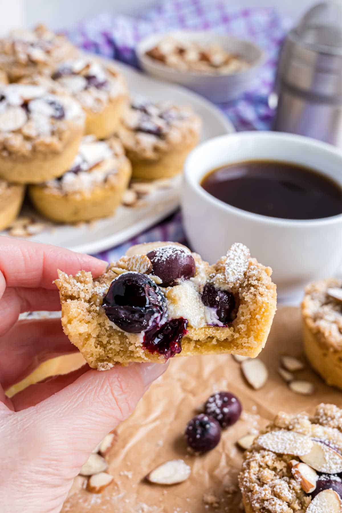 A hand holds a cherry almond pastry with a bite taken out, next to a cup of black coffee and a plate of blueberry coffee cake cookies.