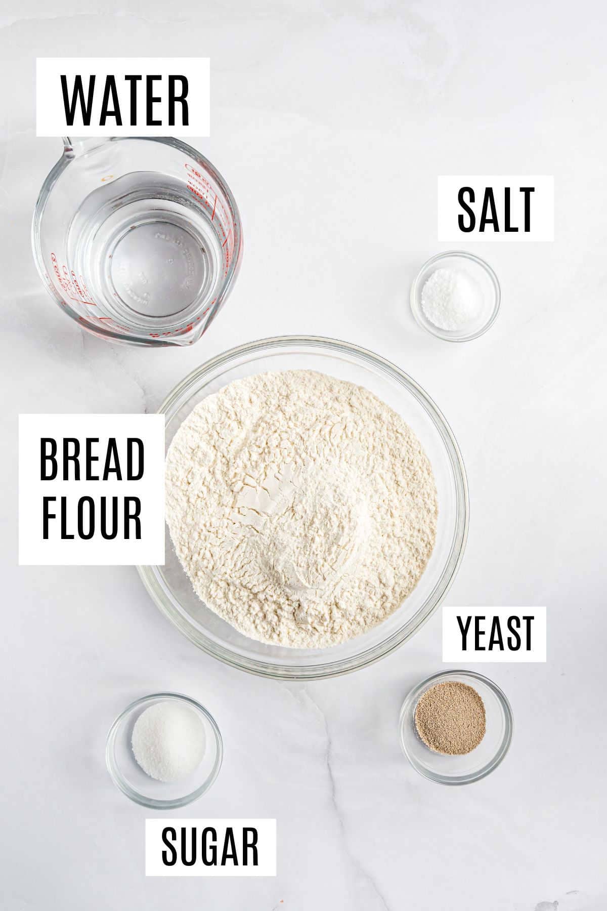 Glass bowls with bread flour, yeast, sugar, salt, and a measuring cup of water on a white surface—perfect for preparing homemade bread bowls.