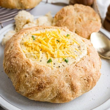 Creamy soup served in a round bread bowl, topped with shredded cheese and herbs, on a white plate—a comforting classic for bread bowl lovers.