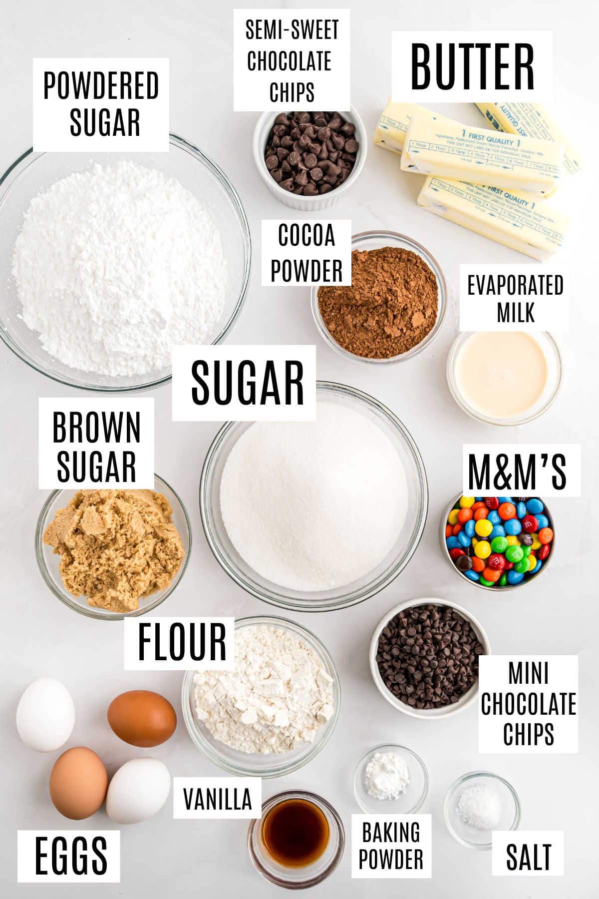 Bowls and containers with labeled baking ingredients on a white surface, including sugar, flour, and chocolate chips.